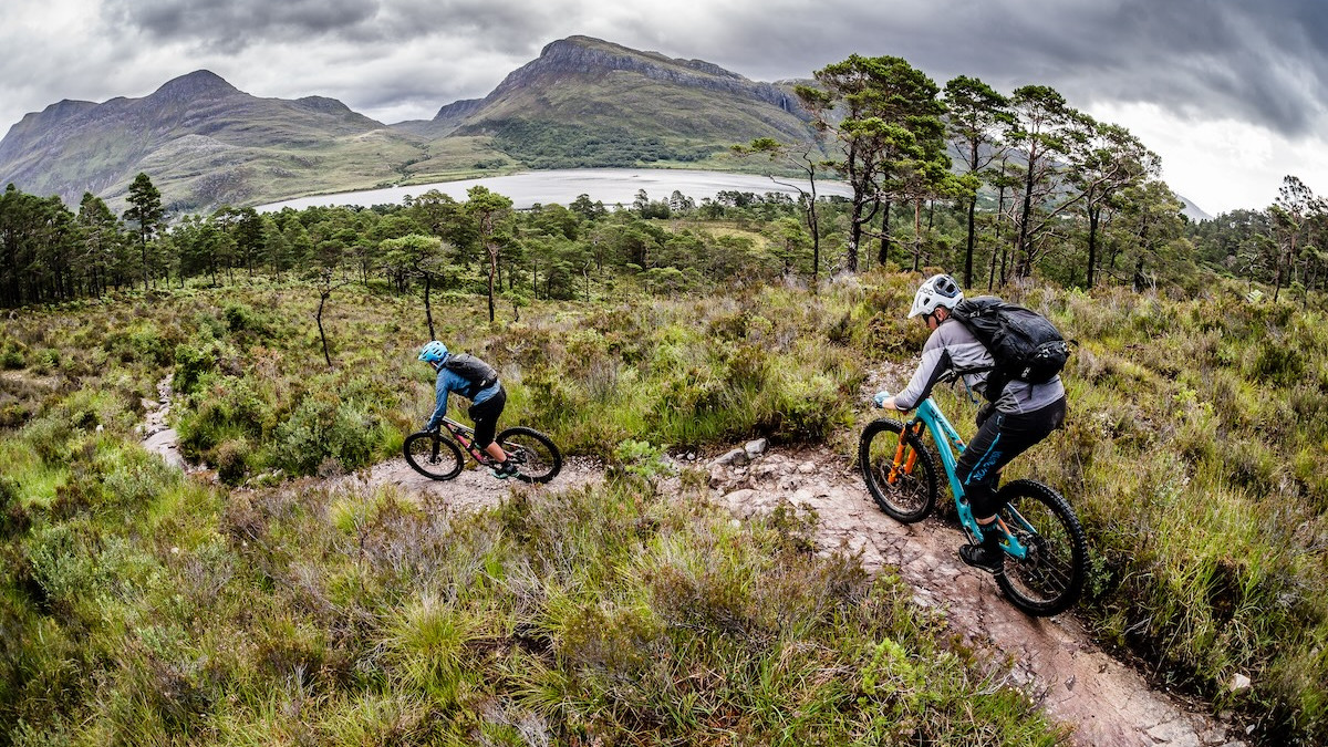 Mountain bikers on rugged trail with scenic backdrop, using self-guided tour tools. Mountain Bike United.