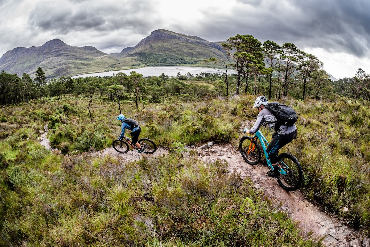 Mountain bikers on rugged trail with scenic backdrop, using self-guided tour tools. Mountain Bike United.